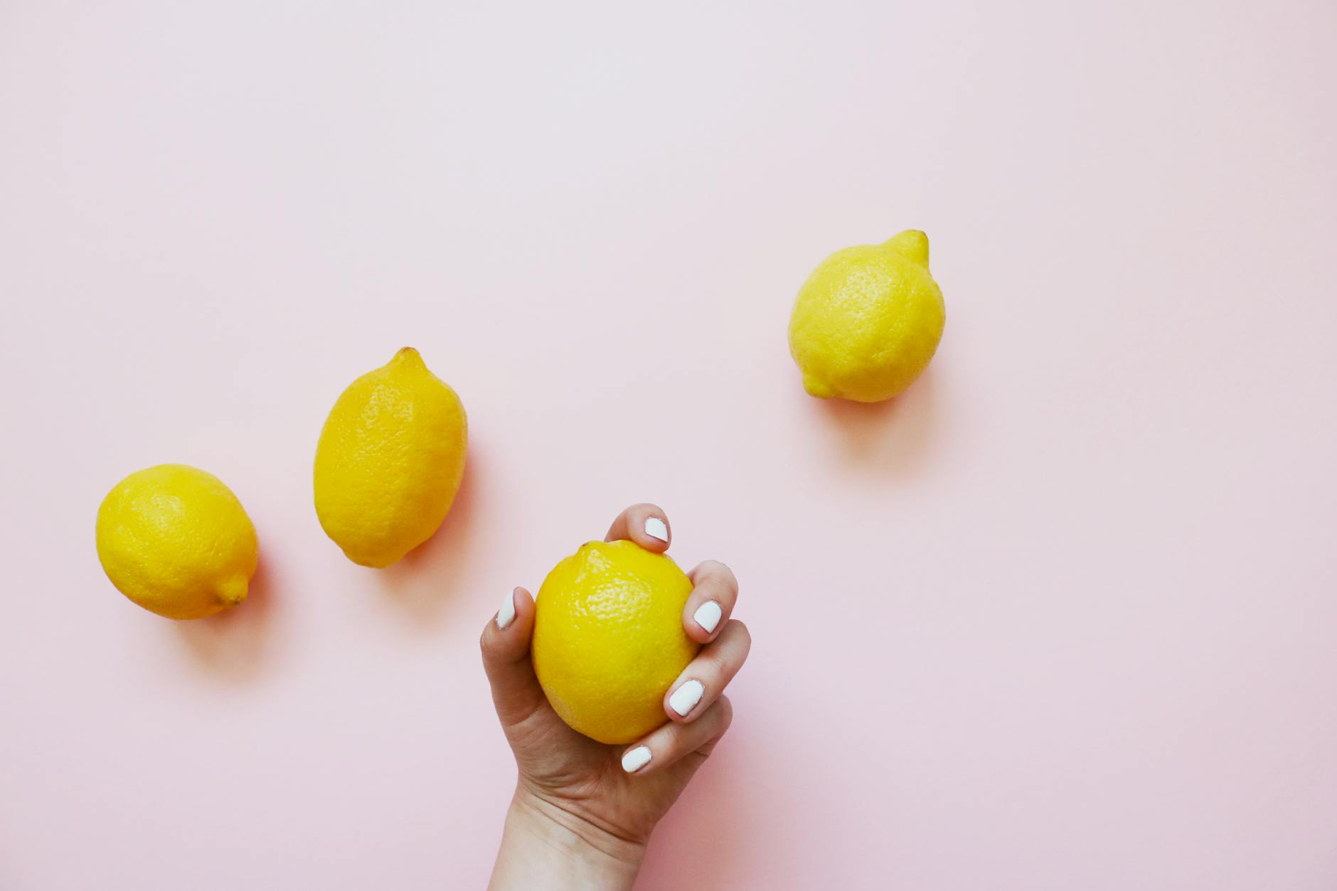A hand holding a fresh lemon on a soft pink background with additional lemons nearby, symbolizing new exploration and citrus-inspired pleasure.